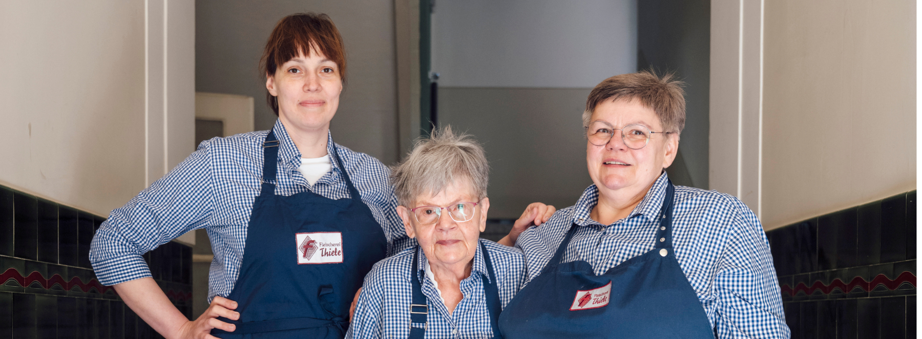 Drei Frauen mit blauen Schürzen mit roter und weißer Aufschrift stehen nebeneinander und lächeln in die Kamera.