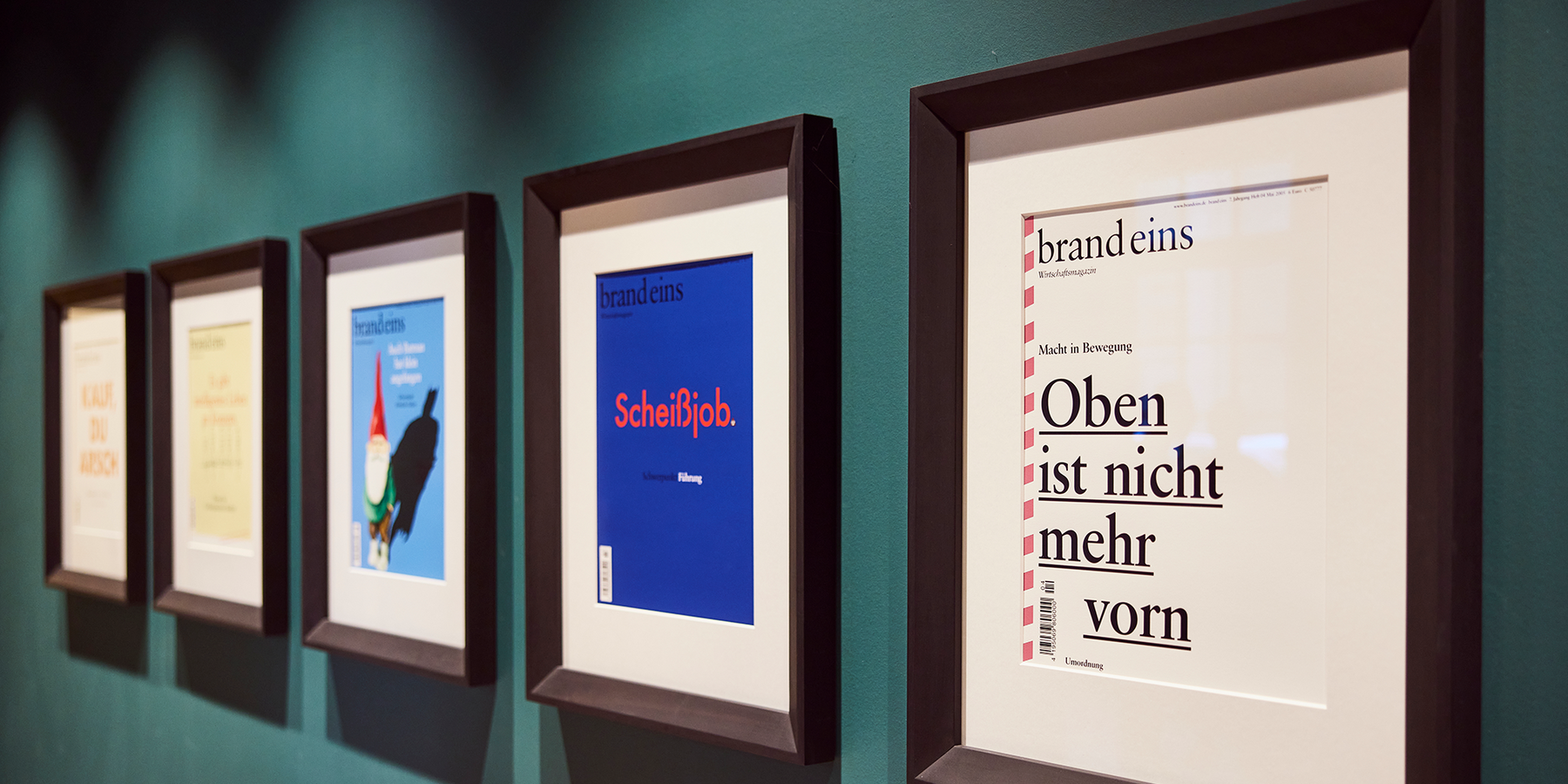 Das Bild zeigt eine Reihe von gerahmten Kunstwerken an einer Wand, wobei der Schwerpunkt auf dem zentralen Kunstwerk liegt, einem Poster mit dem Text "brandeims" und "Schreib". Die anderen Kunstwerke weisen verschiedene Designs und Texte auf, aber das Bild bietet nicht genügend Details, um den spezifischen Inhalt der anderen Werke zu beschreiben. Die Kunstwerke sind an einer Wand aufgehängt, und die Gesamtkomposition lässt auf eine Galerie oder eine Ausstellung schließen.