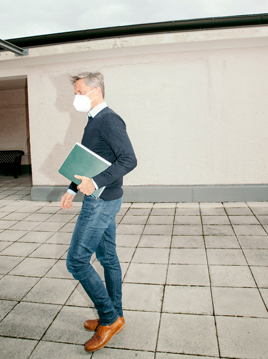 Ein Mann mit einer Gesichtsmaske und einer grünen Mappe in der Hand läuft über einen gefliesten Boden.