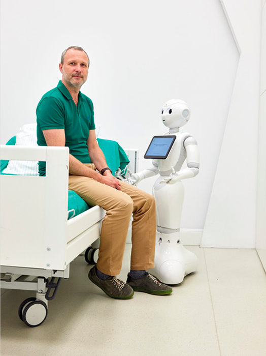 Ein Mann sitzt in einem Krankenhausbett, neben ihm steht ein humanoider Roboter. Der Roboter hat einen Bildschirm auf seiner Brust und scheint mit dem Mann zu interagieren. Die Szene ist in einer Krankenhausumgebung angesiedelt, mit einer weißen Wand und einem Bett im Hintergrund.