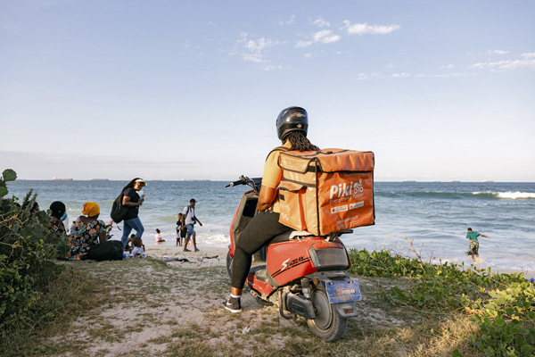 Das Bild zeigt eine Person auf einem roten Motorroller mit einem orangefarbenen Kasten auf dem Rücken, der an einem Sandstrand in der Nähe des Meeres geparkt ist. Im Hintergrund sind mehrere Menschen zu sehen, einige von ihnen stehen, andere sitzen am Strand. Der Himmel ist klar und blau, und das Meer ist im Hintergrund zu sehen.