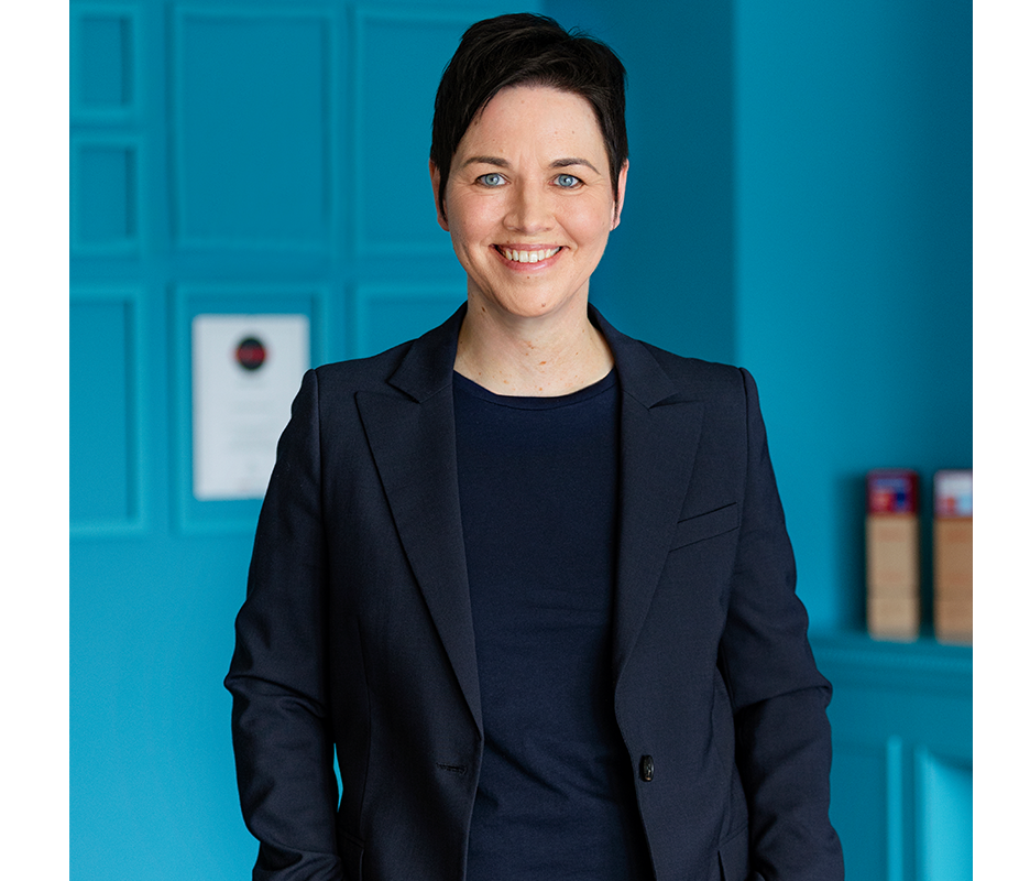 Das Bild zeigt eine Frau, die vor einer blauen Wand steht, lächelt und einen dunkelblauen Blazer trägt. Sie befindet sich in der Mitte des Bildes und hat die Hände in den Taschen. Im Hintergrund sind eine weiße Tür und ein Regal mit verschiedenen Gegenständen zu sehen.