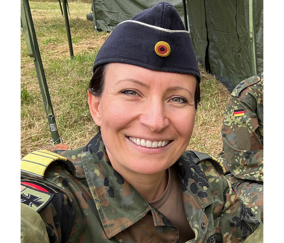 Das Bild zeigt eine lächelnde Frau in einer Militäruniform mit Mütze, die vor einem grünen Zelt steht. Die Frau scheint eine Soldatin zu sein, und ihre Uniform ist mit verschiedenen Abzeichen und Insignien geschmückt. Das Foto ist aus einem niedrigen Winkel aufgenommen, wodurch die Präsenz der Frau und das Zelt im Hintergrund hervorgehoben werden. Die Gesamtkomposition suggeriert eine Szene der Kameradschaft oder des Feierns, möglicherweise im Zusammenhang mit einem militärischen Ereignis oder Anlass.