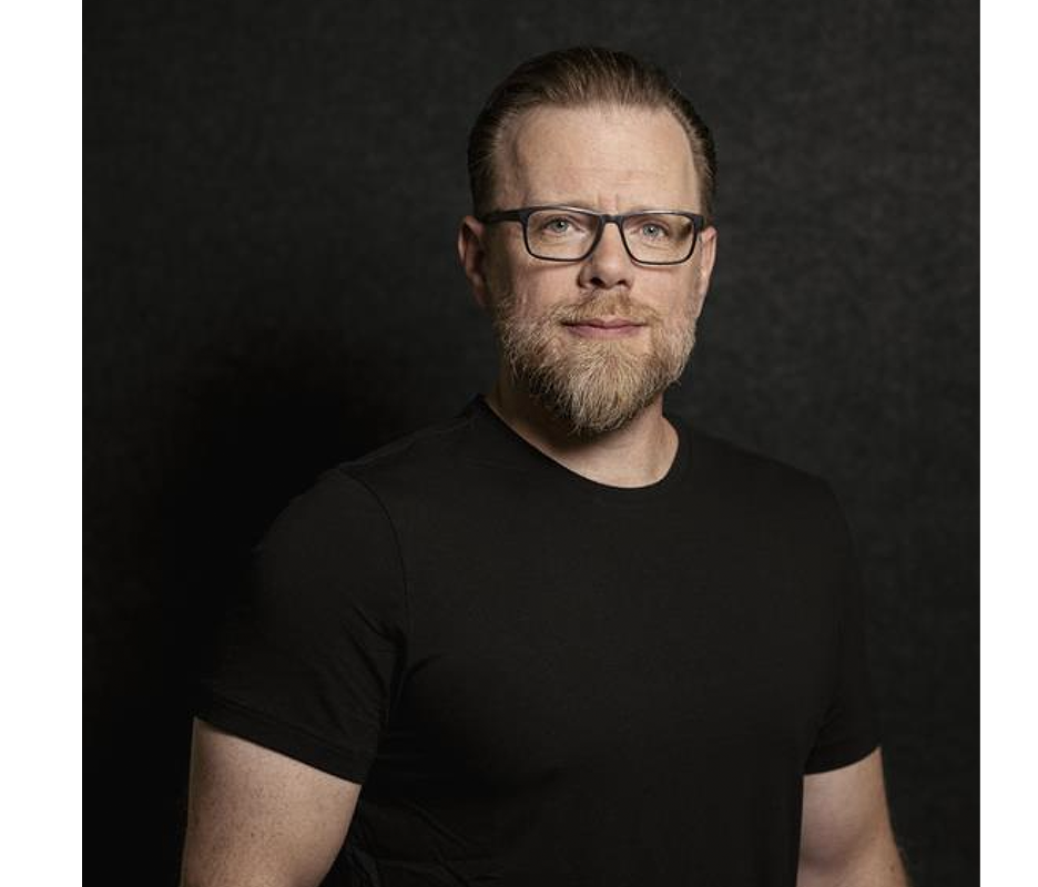 Das Bild zeigt einen Mann mit Bart und Brille, der ein schwarzes T-Shirt trägt und vor einem dunklen Hintergrund steht. Er blickt direkt in die Kamera und hat ein leichtes Lächeln im Gesicht.
