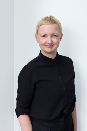 Das Bild zeigt eine Frau mit kurzen blonden Haaren, die ein schwarzes Hemd trägt und vor einer weißen Wand steht. Sie lächelt und schaut direkt in die Kamera, was eine freundliche und zugängliche Ausstrahlung vermittelt. Das Bild wird als eine zugängliche Website beschrieben, die den Leitlinien der EAA (European Association of Advertising Agencies) entspricht, die sicherstellt, dass der Inhalt für alle Nutzer, einschließlich derjenigen mit Behinderungen, leicht zugänglich und navigierbar ist.