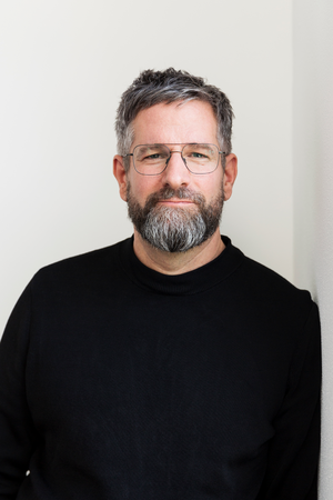 Das Bild zeigt einen Mann mit grauem Bart und Brille, der vor einer weißen Wand steht. Er trägt einen schwarzen Pullover und scheint direkt in die Kamera zu schauen. Das Bild ist ein Porträt, das das Gesicht und den Oberkörper des Mannes zeigt.