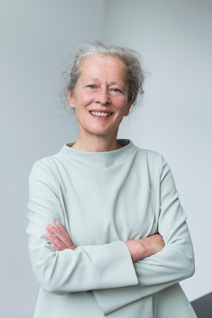 Das Bild zeigt eine lächelnde Frau mit grauem Haar, die vor einem weißen Hintergrund steht. Sie trägt eine helle Bluse und hat die Arme vor sich verschränkt. Die Frau scheint sich in einer professionellen oder formellen Umgebung zu befinden, wie ihre Kleidung und Haltung vermuten lassen.