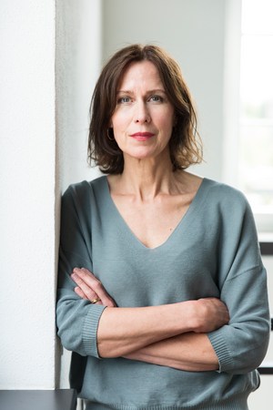 Das Bild zeigt eine Frau, die vor einer weißen Wand steht und die Arme verschränkt. Sie trägt einen grauen Pullover und hat einen neutralen Gesichtsausdruck. Das Foto ist aus einem leicht erhöhten Winkel aufgenommen, der einen klaren Blick auf die Frau und ihre Umgebung ermöglicht.