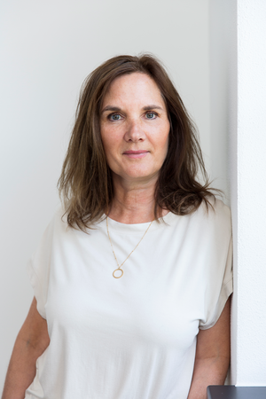 Das Bild zeigt eine Frau mit schulterlangem braunem Haar, die ein weißes Oberteil und eine goldene Halskette trägt. Sie steht vor einer weißen Wand und blickt mit neutralem Blick direkt in die Kamera. Das Bild ist ein Porträt, das das Gesicht und den Oberkörper der Frau zeigt.