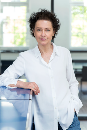 Das Bild zeigt eine Frau, die in einem Raum mit blauer Oberfläche steht und sich gegen eine Wand lehnt. Sie trägt ein weißes Hemd und hat lockiges Haar. Die Frau blickt mit einem Lächeln direkt in die Kamera. Im Hintergrund sind ein Fenster und ein Stuhl zu sehen, was auf eine zwanglose und bequeme Umgebung hindeutet.