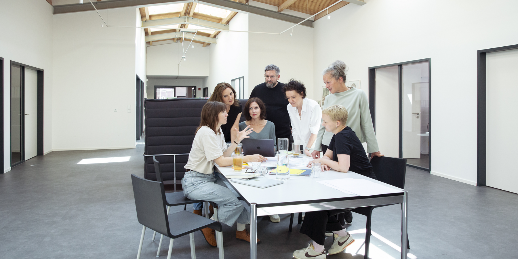 Eine Gruppe von Personen ist in einem modernen Büro um einen Tisch versammelt und führt Gespräche und arbeitet zusammen. Der Tisch ist mit Laptops und anderen Arbeitsmaterialien ausgestattet, was auf ein professionelles Umfeld schließen lässt. Der Raum ist gut beleuchtet, und durch die Fenster fällt natürliches Licht ein, das eine angenehme und einladende Atmosphäre schafft. Die Personen scheinen sich auf die anstehende Aufgabe zu konzentrieren, was auf eine produktive Besprechung oder Brainstorming-Sitzung hindeutet.