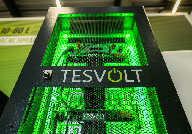 Das Bild zeigt einen Computerturm mit einem grünen netzartigen Gehäuse, das von neongrünen Lichtern beleuchtet wird. Der Turm steht auf einem Tisch, auf dem sich ein Schild mit Text befindet. Der Text auf dem Schild lautet "TESVOLT", was darauf hinweist, dass der Computerturm ein Produkt der Firma "TESVOLT" ist. Das Bild ist für die EAA zugänglich, da es als "Benchmark"- und "Benchmarking"-Bild beschrieben wird, was darauf hindeutet, dass es sich um ein Produkt oder Referenzmaterial im Zusammenhang mit Elektronik oder Computertechnologie handelt.