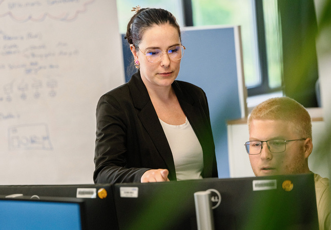 Das Bild zeigt eine Frau in einem schwarzen Blazer und einer Brille, die an einem Schreibtisch steht und auf einen Computerbildschirm schaut, während ein Mann in einem beigen Hemd ebenfalls auf den Bildschirm schaut. Im Hintergrund ist ein Whiteboard mit Schrift zu sehen. Das Bild wird als zugänglich für Menschen mit Behinderungen beschrieben und entspricht den EAA-Richtlinien.