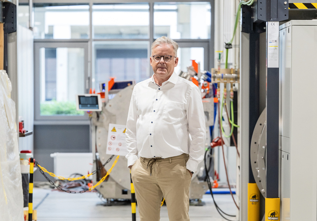 Das Bild zeigt einen Mann, der in einem Raum steht, der mit verschiedenen Geräten und Maschinen gefüllt ist. Er trägt ein weißes Hemd und eine khakifarbene Hose und scheint sich in einer professionellen oder industriellen Umgebung zu befinden. Der Raum ist mit verschiedenen Maschinen und Geräten gefüllt, was auf einen Arbeitsbereich oder eine Fabrikumgebung hindeutet.
