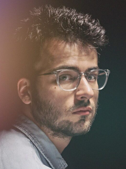 Das Bild zeigt eine Nahaufnahme eines Mannes mit Brille und Bart, der mit ernster Miene direkt in die Kamera schaut. Der Hintergrund ist dunkel, was dazu beiträgt, die Aufmerksamkeit auf das Motiv zu lenken. Zur Kleidung des Mannes gehört ein Hemd mit Kragen, das zur Gesamtkomposition des Bildes beiträgt. Das Bild ist im Hochformat aufgenommen, und der Mann ist die zentrale Figur, ohne dass andere Gegenstände oder Personen zu sehen sind.