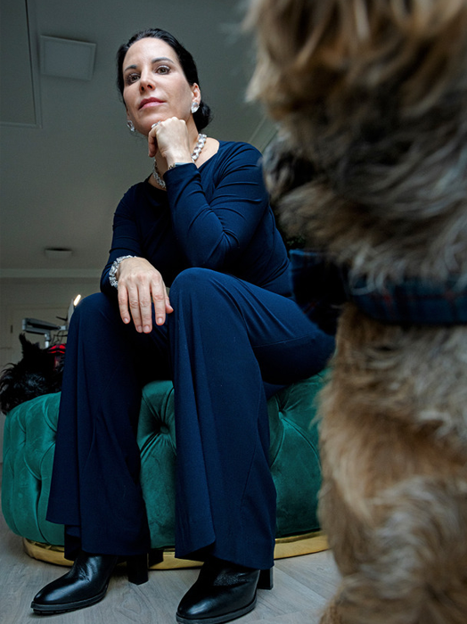 Eine Frau in einem marineblauen Outfit sitzt auf einem grünen Samtsofa, neben ihr steht ein Hund. Die Frau blickt zur Seite, und der Hund scheint sie anzuschauen. Die Szene spielt in einem Raum mit einem Holzboden und einer weißen Wand im Hintergrund.
