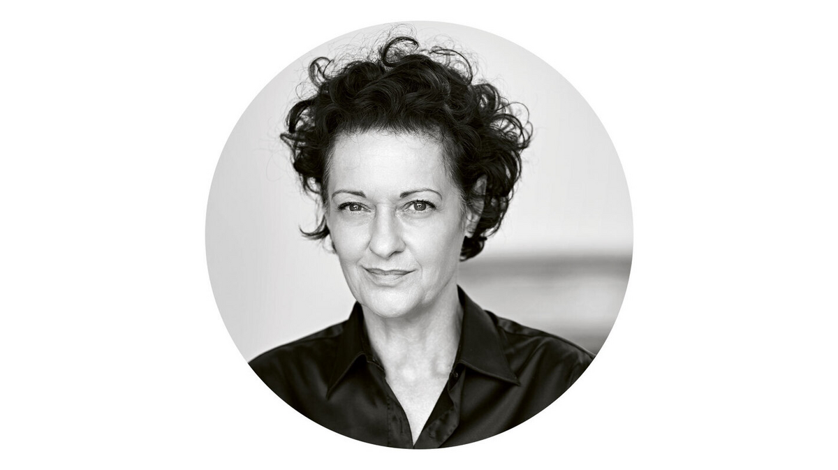 Das Bild ist ein Schwarz-Weiß-Porträt einer Frau mit lockigem Haar, die mit einem leichten Lächeln direkt in die Kamera schaut. Das Porträt wird von einem kreisförmigen Rahmen eingerahmt, der ihm ein professionelles und gepflegtes Aussehen verleiht. Die Frau ist mit einem schwarzen Hemd bekleidet, das einen Kontrast zu den monochromen Farbtönen des Bildes bildet. Die Gesamtkomposition und die Verwendung einer einfachen Farbpalette verleihen dem Porträt eine zeitlose und klassische Ausstrahlung.