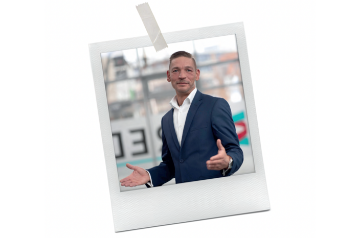 Das Bild zeigt einen Mann im Anzug, der vor einem unscharfen Hintergrund steht, möglicherweise eine Straße in der Stadt. Er trägt ein weißes Hemd und eine blaue Jacke, und er scheint mit den Händen zu gestikulieren. Das Bild ist von einem weißen Rand umrahmt, und in der linken oberen Ecke befindet sich ein Stück Klebeband. Die Gesamtkomposition lässt auf einen beruflichen oder geschäftlichen Kontext schließen, aber die spezifischen Details des Bildes sind aufgrund der Unschärfe und des fehlenden Kontexts nicht klar.