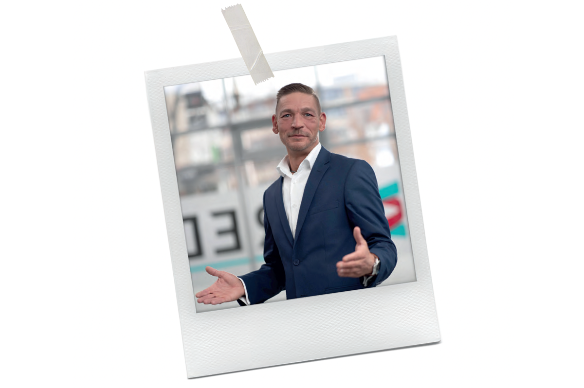 Das Bild zeigt einen Mann im Anzug, der vor einem unscharfen Hintergrund steht, möglicherweise eine Straße in der Stadt. Er trägt ein weißes Hemd und eine blaue Jacke, und er scheint mit den Händen zu gestikulieren. Das Bild ist von einem weißen Rand umrahmt, und in der linken oberen Ecke befindet sich ein Stück Klebeband. Die Gesamtkomposition lässt auf einen beruflichen oder geschäftlichen Kontext schließen, aber die spezifischen Details des Bildes sind aufgrund der Unschärfe und des fehlenden Kontexts nicht klar.