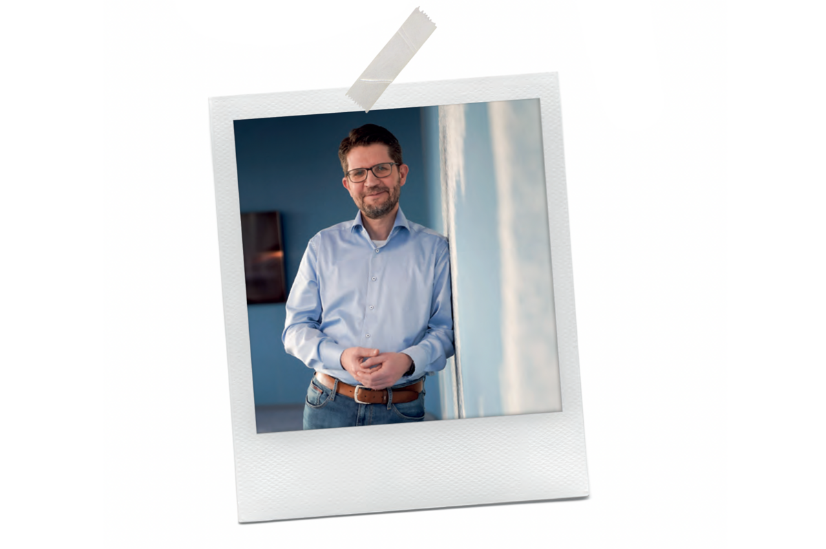 Bei dem Bild handelt es sich um ein Polaroid-Foto eines Mannes, der lächelnd vor einem Fenster steht und ein blaues Hemd und eine Brille trägt. Das Foto ist von einem weißen Rahmen umgeben, und in der oberen linken Ecke des Rahmens befindet sich ein Stück Klebeband. Das Bild wird als für alle Nutzer zugänglich beschrieben, und es wird als Screenshot einer Website beschrieben, die den Richtlinien der EAA (European Association of Advertising Agencies) entspricht.