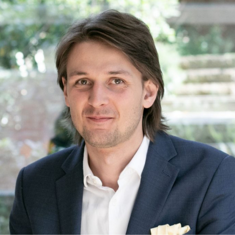 Das Bild zeigt einen Mann mit langen Haaren, der eine dunkelblaue Anzugsjacke und ein weißes Hemd trägt. Er lächelt in die Kamera, der Hintergrund ist unscharf und scheint ein Garten oder ein Außenbereich zu sein.