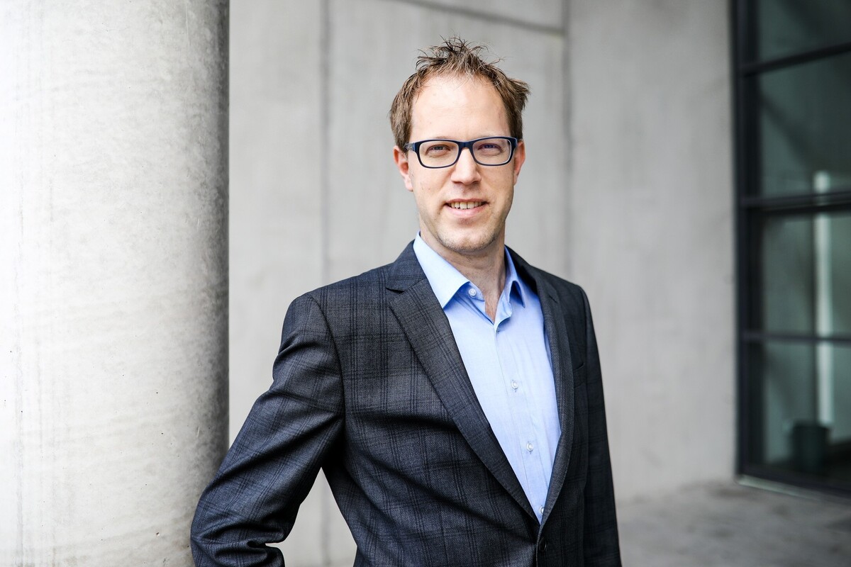 Das Bild zeigt einen Mann in einem Anzug und mit Brille, der vor einem grauen Gebäude steht. Er lächelt und scheint für ein Porträt zu posieren. Der Mann trägt ein blaues Hemd und eine dunkelgraue Anzugsjacke. Der Hintergrund des Bildes ist ein graues Gebäude, das einen neutralen Hintergrund für die Kleidung des Mannes bietet. Das Bild wird als für alle Nutzer zugänglich beschrieben, auch für Menschen mit Sehbehinderungen, da es den EAA-Richtlinien entspricht.