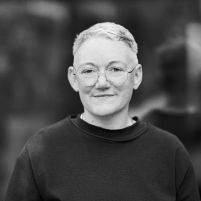 Das Bild ist ein Schwarz-Weiß-Porträt einer lächelnden Frau mit kurzen Haaren und Brille. Sie trägt einen dunklen Pullover und scheint das Hauptmotiv des Bildes zu sein. Der Hintergrund ist unscharf und lenkt die Aufmerksamkeit auf die Frau und ihren Ausdruck. Das Bild ist im Hochformat gehalten, und die Frau steht im Mittelpunkt, ohne dass andere Objekte oder Text vorhanden sind.
