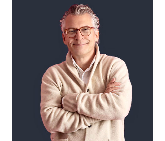 Das Bild zeigt einen lächelnden Mann mit grauem Haar und Brille, der vor einem dunklen Hintergrund steht. Er trägt eine cremefarbene Strickjacke und hat die Arme vor sich verschränkt. Der Mann scheint eine entspannte und zuversichtliche Pose einzunehmen, mit einem warmen und freundlichen Ausdruck.