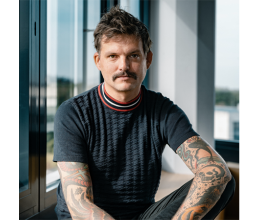Das Bild zeigt einen Mann mit Schnurrbart und Tattoos, der vor einem Fenster sitzt. Er trägt ein schwarzes Hemd mit einem rot-weiß gestreiften Kragen. Der Mann blickt direkt in die Kamera, so dass seine Gesichtszüge deutlich zu erkennen sind. Im Hintergrund des Bildes ist die Ansicht eines Gebäudes zu sehen, was darauf hindeutet, dass sich der Mann in einem öffentlichen oder halböffentlichen Raum aufhalten könnte.