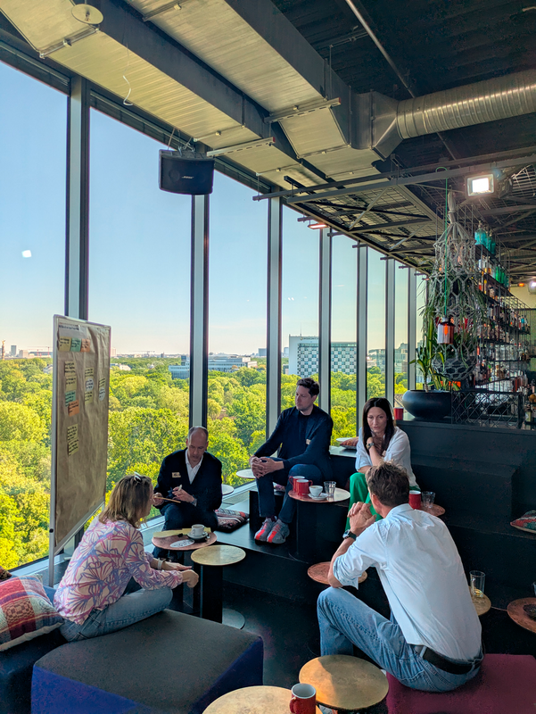 Das Bild zeigt eine Gruppe von Menschen, die in einem modernen Großraumbüro mit großen Fenstern und Blick auf eine Stadtlandschaft um einen Tisch sitzen. Das Büro hat ein schlichtes, modernes Design mit schwarzer und weißer Farbgebung. Die Personen scheinen in eine Diskussion oder Besprechung vertieft zu sein, wobei einige von ihnen eine Tasse Kaffee oder Tee in der Hand halten. Der Büroraum ist mit schwarzen Tischen und Stühlen ausgestattet, und Topfpflanzen und andere dekorative Elemente tragen zum Ambiente bei.