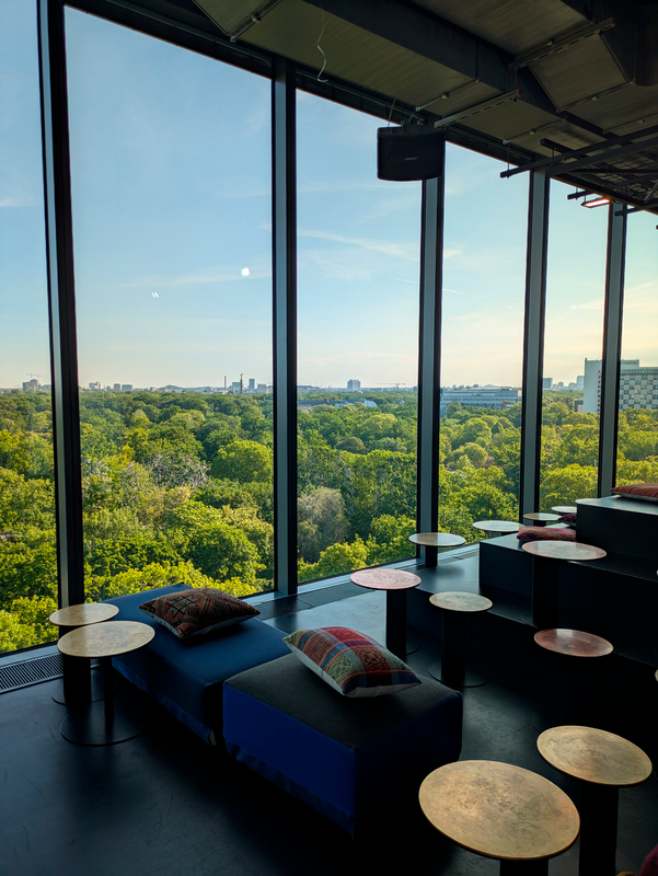 Das Bild zeigt ein modernes Großraumbüro mit großen Fenstern, die einen Panoramablick auf einen üppigen, grünen Wald bieten. Das Büro ist mit verschiedenen Sitzgelegenheiten ausgestattet, darunter runde Tische und Sofas, die so angeordnet sind, dass sie die Zusammenarbeit und Interaktion zwischen den Mitarbeitern fördern. Das Gesamtdesign des Raums ist schlicht und modern, mit dem Schwerpunkt auf natürlichem Licht und einer Verbindung zur Natur.
