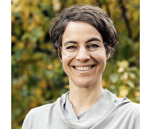 Das Bild zeigt eine lächelnde Frau mit kurzen Haaren und Brille, die ein graues Oberteil trägt und vor einem verschwommenen grünen Hintergrund steht.