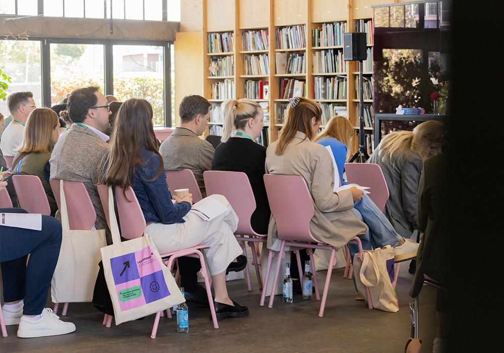 Das Bild zeigt eine Gruppe von Personen, die auf Stühlen in einer Bibliothek oder einem Konferenzraum sitzen, mit Bücherregalen im Hintergrund.