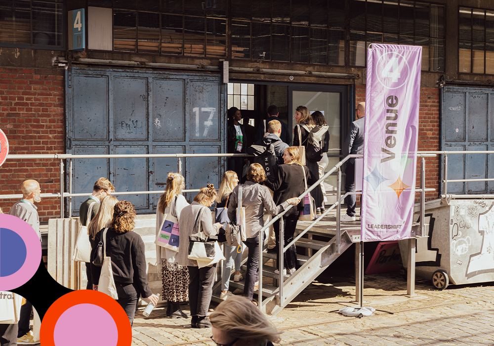 Das Bild zeigt eine Gruppe von Menschen, die auf einer Treppe vor einem Gebäude stehen, wobei einige von ihnen Taschen tragen. In der Szene ist ein lila Banner mit dem Wort "LEADERSHIP" zu sehen. Das Bild scheint aus einem niedrigen Winkel aufgenommen worden zu sein, wodurch die Höhe der Treppe und die Statur der Personen betont werden. Die Gesamtszene deutet auf einen öffentlichen oder kommerziellen Rahmen hin, möglicherweise einen Veranstaltungsort oder eine Veranstaltungsfläche.