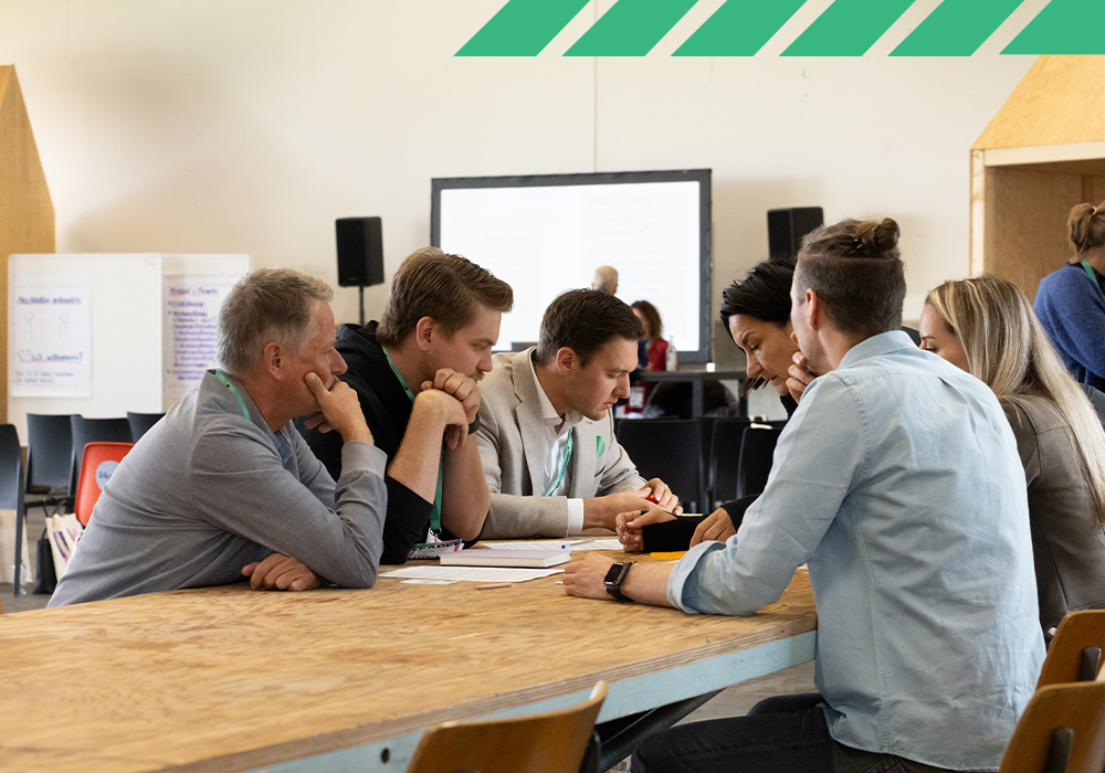 Das Bild zeigt eine Gruppe von Personen, die an einem Tisch sitzen und miteinander diskutieren und zusammenarbeiten. Es sind mehrere Personen zu sehen, von denen einige auf Stühlen sitzen und andere stehen. Der Tisch ist mit Papieren und Dokumenten bedeckt, was auf eine Sitzung oder Diskussion hindeutet. Bei dem Raum scheint es sich um einen Konferenz- oder Besprechungsraum zu handeln, in dem im Hintergrund ein großer Bildschirm oder Projektor zu sehen ist.