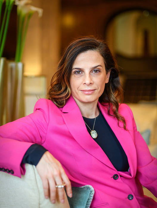 Das Bild zeigt eine Frau mit langen braunen Haaren, die einen hellrosa Blazer trägt, auf einer Couch sitzt und in die Kamera lächelt. Sie befindet sich in der Mitte des Bildes, und im Hintergrund ist eine Topfpflanze zu sehen. Das Bild scheint ein Porträt zu sein, wobei der Schwerpunkt auf dem Ausdruck und der Kleidung der Frau liegt.