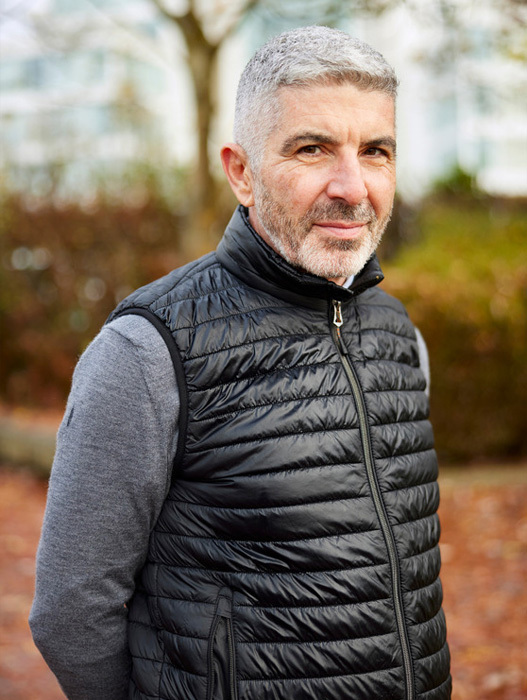 Das Bild zeigt einen Mann mit grauem Haar und Bart, der in einem Park steht. Er trägt eine schwarze Pufferweste und einen grauen Pullover. Der Mann blickt direkt in die Kamera, so dass sein Gesicht klar zu erkennen ist. Der Hintergrund des Bildes ist unscharf und suggeriert eine Parklandschaft mit Bäumen und Laub.