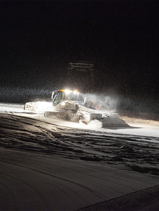 Das Bild zeigt einen Schneepflug, der nachts einen Weg durch eine verschneite Landschaft räumt. Der Pflug räumt aktiv den Schnee und hinterlässt eine Spur. Die Szene wird durch die hellen Lichter des Pfluges beleuchtet, die im Vordergrund zu sehen sind. Der Hintergrund ist dunkel und hebt den schneebedeckten Boden und die Bewegung des Pfluges hervor. Das Bild ist in Farbe und bietet einen lebhaften Kontrast zum weißen Schnee und den Lichtern des Pfluges.
