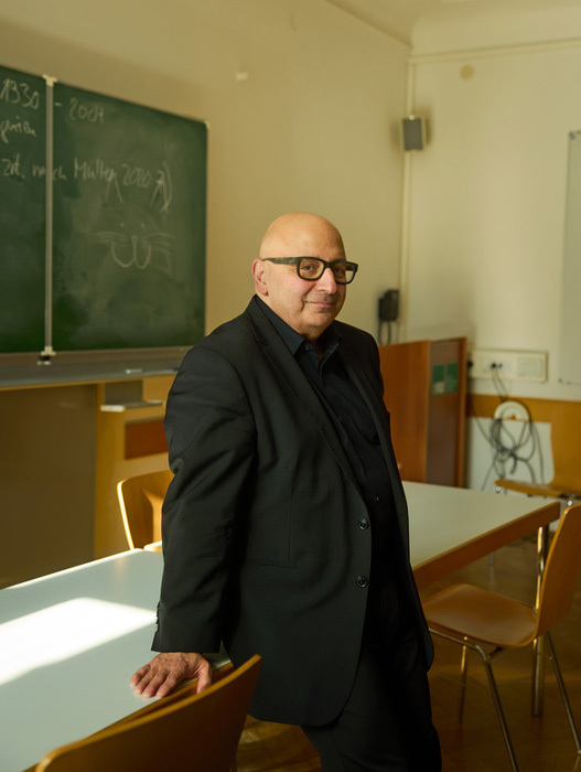 Das Bild zeigt einen Mann, der in einem Klassenzimmer steht und sich mit der Hand auf einen Tisch stützt. Er trägt einen schwarzen Anzug und eine Brille, und im Hintergrund ist eine Kreidetafel zu sehen. Das Klassenzimmer scheint gut ausgestattet zu sein, mit Stühlen und einem Schreibtisch.