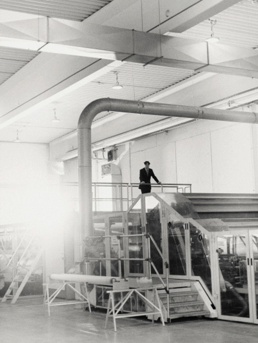 Bei dem Bild handelt es sich um eine Schwarz-Weiß-Fotografie einer industriellen Umgebung, die einen Mann zeigt, der auf einer Plattform oder einer erhöhten Struktur innerhalb eines großen Lagerhauses steht. Der Mann scheint die Umgebung zu beobachten oder möglicherweise den Betrieb der Maschinen zu überwachen. Das Foto ist aus einem niedrigen Winkel aufgenommen, wodurch die Höhe der Plattform und die Position des Mannes betont werden. Im Hintergrund sind eine Reihe von Rohren und andere industrielle Geräte zu sehen, die auf einen gut ausgestatteten Arbeitsbereich hindeuten.