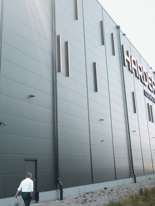 Das Bild zeigt einen Mann, der an einem großen, modernen grauen Gebäude mit vertikalen Linien an der Fassade vorbeigeht. Das Gebäude trägt ein auffälliges "HARPER"-Schild an der Seite. Die Szene scheint in einer städtischen Umgebung angesiedelt zu sein, wobei der Mann auf dem Gehweg vor dem Gebäude geht.