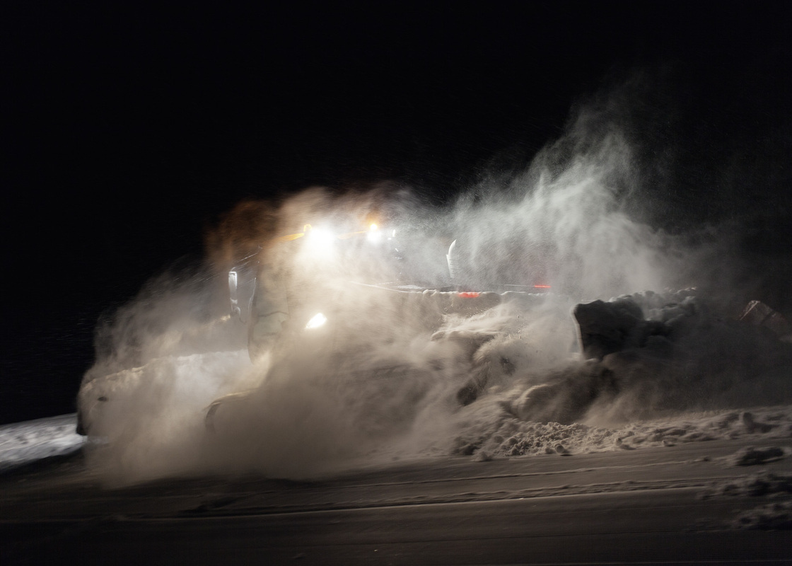 Das Bild zeigt einen Schneepflug, der nachts einen Weg durch eine verschneite Landschaft räumt. Der Pflug räumt aktiv den Schnee und erzeugt dabei eine große Schneeschaufel. Die Szene wird von den hellen Lichtern des Pfluges beleuchtet, die in der oberen linken Ecke des Bildes zu sehen sind. Die Gesamtatmosphäre vermittelt ein Gefühl von Aktivität und Fortschritt inmitten der winterlichen Bedingungen.