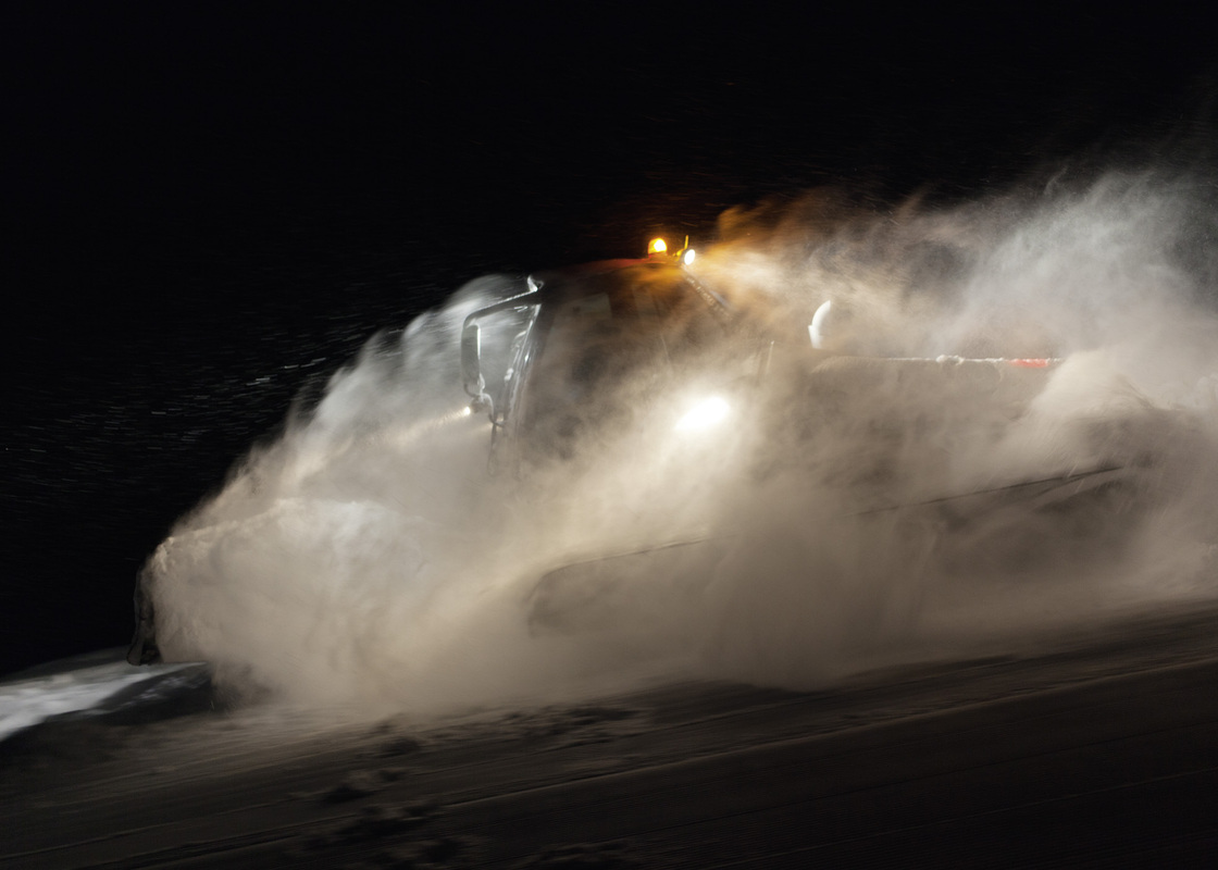 Das Bild zeigt einen Schneepflug in Aktion, der nachts einen Weg durch eine verschneite Landschaft freiräumt. Der Pflug räumt aktiv den Schnee von der Straße und erzeugt dabei eine große Schneeschaufel. Die Szene wird von den hellen Lichtern des Pfluges beleuchtet, die in der oberen rechten Ecke des Bildes zu sehen sind. Die Gesamtkomposition suggeriert ein Gefühl der Bewegung und des Fortschritts, während der Schneepflug seine Arbeit fortsetzt.