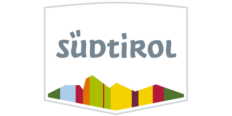 Das Bild zeigt ein Logo mit einem schildförmigen Design und einer Bergkette im Hintergrund. Die Gebirgskette besteht aus verschiedenfarbigen Gebäuden, die eine lebendige und visuell ansprechende Szene bilden. Der Schriftzug "südtirol" ist prominent in der Mitte des Schildes angebracht und steht wahrscheinlich für den Namen oder Slogan des Unternehmens oder der Organisation, die mit dem Logo verbunden ist. Das Gesamtdesign ist einfach und doch auffällig, was es zu einem zugänglichen und effektiven Logo für eine Website macht.