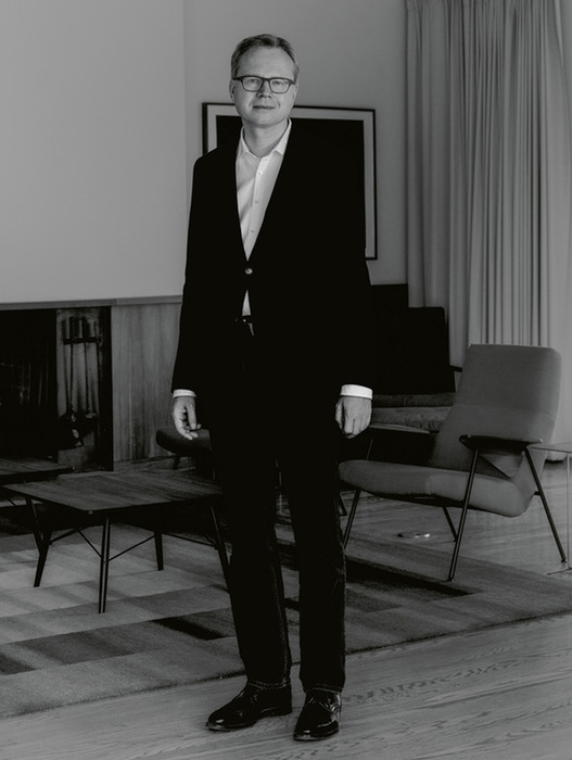 Das Bild ist eine Schwarz-Weiß-Fotografie eines Mannes, der in einem Raum steht. Er ist mit Anzug und Krawatte bekleidet und steht vor einem Kamin. Der Raum ist mit einer Couch, einem Stuhl und einem Couchtisch ausgestattet. Der Mann scheint das Hauptmotiv des Fotos zu sein, und die Gesamtkomposition lässt auf eine formale oder professionelle Umgebung schließen.