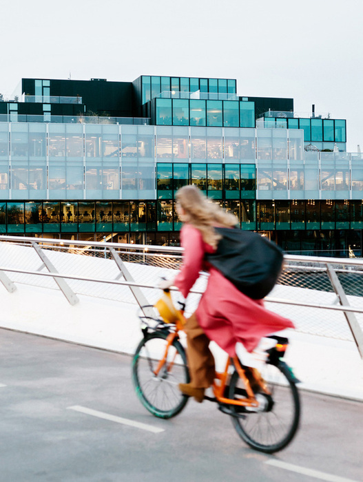 Das Bild zeigt eine Frau, die mit einem orangefarbenen Fahrrad auf einer Brücke fährt, mit einem modernen Glasgebäude im Hintergrund. Die Szene ist in Bewegung eingefangen, was ein Gefühl von Geschwindigkeit und Bewegung vermittelt. Die Frau trägt eine rosafarbene Jacke und eine schwarze Tasche, die der Szene einen Farbakzent verleiht. Das Bild ist aus einem niedrigen Winkel aufgenommen, wodurch die Höhe der Brücke und die Position der Frau auf dem Fahrrad betont werden. Die Gesamtkomposition ist visuell ansprechend und fängt die Essenz des städtischen Lebens ein.