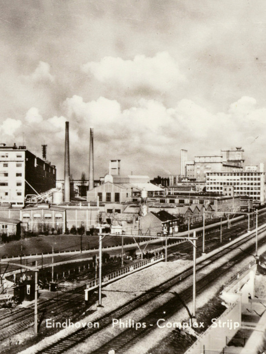 Bei dem Bild handelt es sich um eine Schwarz-Weiß-Aufnahme einer Stadtlandschaft mit Industriegebäuden, einer Eisenbahnstrecke und einem bewölkten Himmel. Das Foto scheint von einem hohen Aussichtspunkt aus aufgenommen worden zu sein und bietet einen umfassenden Überblick über das Layout und die Infrastruktur der Stadt. Das Bild ist am unteren Rand mit dem Text "Eindhaven Philips Complex Stripp" beschriftet, was auf den Standort und den Namen des Fotografen hinweist. Das Foto wird als altes Foto beschrieben, was darauf hindeutet, dass es eine historische Bedeutung haben oder ein wertvolles historisches Dokument sein könnte.