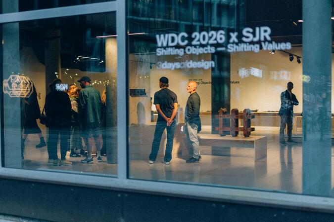 Das Bild zeigt eine Gruppe von Menschen, die sich in einem Schaufenster versammelt haben, in dem ein großes Glasfenster den Text "WDC 2026 x SJR Shifting Objects - Shifting Values 3 days of design" und ein Logo zeigt. Das Geschäft scheint eine Kunstgalerie oder ein Ausstellungsraum zu sein, mit verschiedenen Kunstwerken und Skulpturen, die durch das Fenster sichtbar sind.