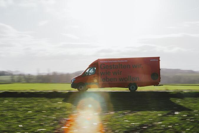 Das Bild zeigt einen roten Lieferwagen, der auf einer Straße durch ein grasbewachsenes Feld fährt. Auf der Seite des Lieferwagens ist eine große Werbung angebracht, und es ist der Text "Gestalten wir, wie wir leben wollen" zu sehen, was so viel bedeutet wie "Die Welt verändert sich, wir müssen uns anpassen." Der Lieferwagen bewegt sich auf die rechte Seite des Bildes zu, und der Hintergrund zeigt einen bewölkten Himmel.