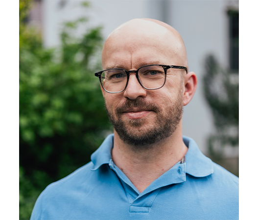 Das Bild zeigt einen Mann mit Brille und blauem Poloshirt, der im Freien vor einem unscharfen Hintergrund steht.